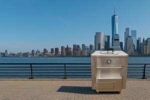 Stainless steel tandoor oven placed outdoors by the New Jersey waterfront with the New York City skyline visible across the Hudson River under a clear blue sky.