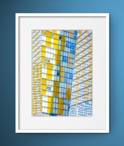 A framed fine art photograph of a modern glass building with yellow and blue reflective panels, showcasing Dina Marie’s exploration of structure, color, and light.