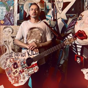 Self-portrait of independent musician Scooter Scudieri in his Shepherdstown studio holding his hand-painted guitar covered in stickers and artwork, featuring the phrase “This Machine Does Not Kill,” a nod to creative resistance and artistic freedom.