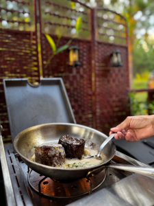 Close-up of a chef’s hand spooning sauce over two pieces of seared beef tenderloin in a pan on an outdoor grill, with a rustic privacy screen and forest backdrop.