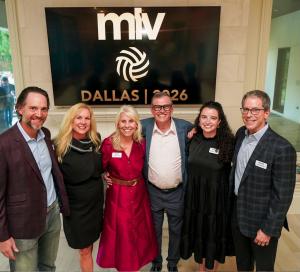 Six people stand smiling in front of a large screen displaying the Major League Volleyball (MLV) Dallas 2026 logo. The group includes three men and three women dressed in business and cocktail attire, posing arm-in-arm in a warmly lit event space. The ima