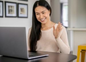 A smiling woman waving during a virtual therapy session on her laptop in a home environment.