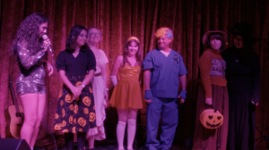 Farrah Mechael stands on stage with Halloween-costumed guests at Universal Bar & Grill under pink-purple lights during her Halloween Eve performance.