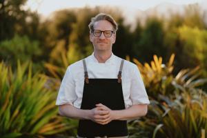 Toby Stuart standing outdoors at Domu Retreat with panoramic views of the Abel Tasman coast behind him, wearing a relaxed chef’s jacket and smiling beside natural timber architecture.