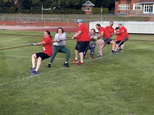 A photo from HM Land Registry's sports day showing 8 people all pulling on a rope in a tug-o-war on the edge of a grass sports pitch with a pavillion in the background.