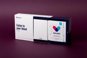 A white box is shown, pulled open. The cover of the box shows the Thriva logo, and the box also contains the instructions for the device within it, which is a blood collection device for use at home.