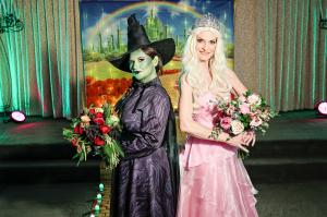 A woman dressed as Elphaba in black with a green face and witches hat holds a large bouquet with dark flowers next to a woman dressed as Glinda in light pink with a crown holds a bouquet of light pinks and greens as they stand on a yellow brick road insid