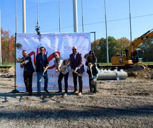 Three men and one woman stand in front of a step and repeat with shovels throwing dirt to symbolize a groundbreaking for a convenience store.