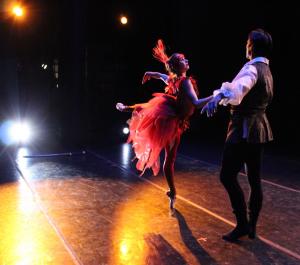 Two ballet dancers performing on stage under dramatic lighting: a female-presenting dancer in a vibrant red feathered costume balanced en pointe, and a male-presenting dancer in a dark period-style outfit reaching toward her.