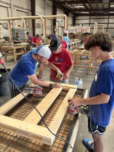 Volunteers wearing SHP AZ-Phoenix shirts work together in a large workshop to sand and assemble wooden bed frames, with multiple bed-building stations visible throughout the space and a "WorkSHoP" sign on the wall.