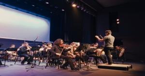 Composer Jonathan Corbie conducts a live orchestra on stage, surrounded by musicians playing strings and brass instruments