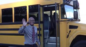 Superintendent Dr. Anton Anthony smiling and waving beside a yellow school bus after helping with morning transportation duties.
