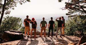 A group of men stand at the edge of an overlook, looking out over the hillside.