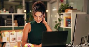 Young woman rubbing her neck while using a computer, illustrating “tech neck” and poor posture from prolonged screen time.