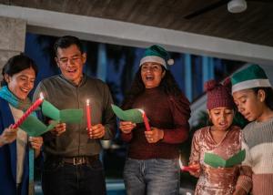 Multi-generational family singing holiday carols together indoors, holding green songbooks and candles, wearing festive holiday attire including Santa hats, demonstrating family bonding and authentic holiday connection.