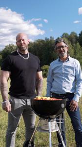 Two men standing by a barbecue grill in a sunny field, showing how Grabbterapi (Dude Therapy) uses everyday activities to encourage open conversation.