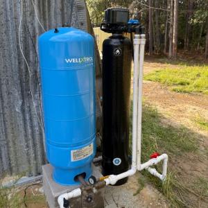Outdoor well system with a blue WellXtrol pressure tank and a black filtration unit connected by PVC piping, installed beside a metal structure near a wooded area.
