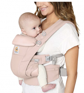 A smiling woman in a white t-shirt carries a baby in a blush pink Ergobaby carrier, holding the child close to her chest with both facing inward against a plain background.