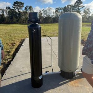 Outdoor well system featuring a tall black filtration unit and a large gray pressure tank connected by PVC piping, set on a concrete pad in an open grassy area