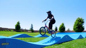 A person wearing a helmet rides a bike over a blue modular pump track, lifting off one of the rollers in a grassy outdoor area.