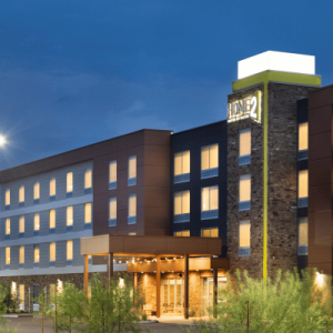 Exterior view of a modern Home2 Suites hotel, featuring multiple stories, bright windows, and a welcoming entrance under a twilight sky.