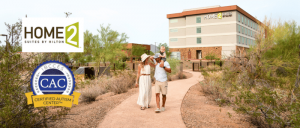 A family strolls on a path near Home2 Suites by Hilton, showcasing the Certified Autism Center emblem and a serene desert landscape.