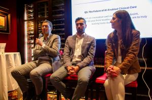 Three healthcare professionals seated on a panel; the person on the left speaks into a microphone while the others listen, with a presentation slide about AI-enabled clinical care displayed behind them.