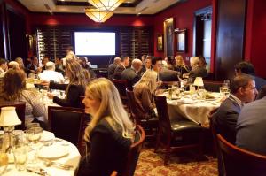 Crowded dining room filled with healthcare professionals seated at round tables, talking and eating during a formal dinner event, with a presentation screen visible in the background.