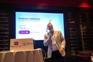 A man in a blazer stands onstage holding a microphone and speaking in front of a large screen displaying Predictive Healthcare’s presentation, discussing how the MyHealthPal platform improves clinical workflows and helps reduce surgical site infections.