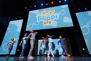 Performers in colorful outfits energetically dance on stage in front of a large screen displaying the "Blippi" logo. The atmosphere is lively and fun.