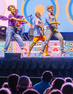 Three performers dance energetically on stage with colorful backgrounds and lights. They wear vibrant costumes, creating a lively and joyful atmosphere.