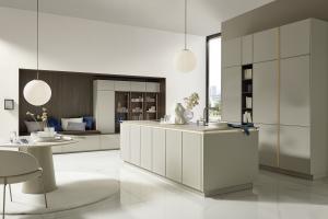Open plan kitchen with central light beige island, tall cabinets, built in bench and shelving niche in dark wood, large window and white marble floor.