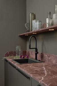 Close up of pink marble countertop and backsplash with black sink and faucet, glass vases and under shelf lights against a taupe wall.