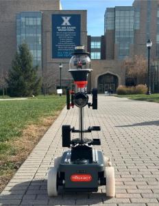 TASKBOT SCOUT, a compact autonomous robot, patrols a brick pathway in front of Xavier University’s main academic building, with its advanced camera and sensor system prominently visible.