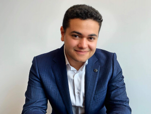Professional headshot of a young man (Michael Black) wearing a navy blue patterned suit jacket, white shirt and a lapel pin, smiling against a white background.