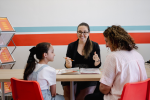 A female tutor or advisor smiles while consulting with a young girl and her mother across a table in a brightly colored office.