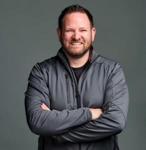 Professional headshot of a smiling man with short hair and a trimmed beard, wearing a gray zip-up jacket and standing with his arms crossed against a plain gray background.