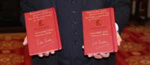 Photograph of Two Guildhawk Queens' Award Commemorative Notebooks in Red with the Guildhawk Girl Registered Trademark Symbol and Signed in Silver Ink by The Right Honourable Peter Estlin The Lord Mayor of London . Books being held by David Clark wearing a