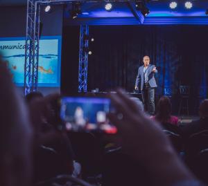 Dr. Dennis Cummins delivering a keynote on stage with an audience and live recording in view.