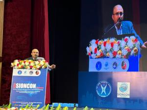 Dr. Mohana Rao Patibandla speaking at the podium during SIONCON 2025 at NIMHANS Bengaluru, presenting intraoperative neuromonitoring research with his talk displayed on a large screen beside him.
