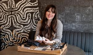 Ariana Anderson, Founder and Principal Designer of Ariana Designs & Interiors, smiles while showcasing design material samples and fabric swatches at a creative workspace with patterned wallpaper backdrop.