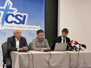 A translator sits between two male speakers at a press conference