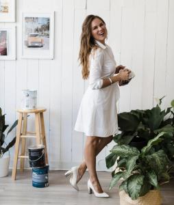 ARIID Build & Remodel professional woman smiling in creative workspace with paint cans, mood boards, and design samples, representing the design-build expertise offered by the firm.