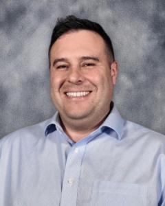 Professional headshot of Dr. Andrew Van Scoyk, a Family Medicine physician, smiling while wearing a light blue collared shirt against a gray textured background.