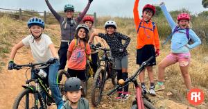 Ride Krew coach standing with a group of smiling young riders during a youth mountain biking program, showing kids learning skills and building confidence on the trail.