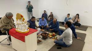 A group of people attending a Ganesh puja ceremony during the inauguration of a new office space, with a priest performing rituals and participants seated on mats around the ceremonial setup.