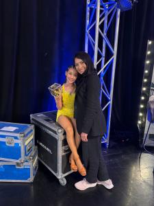 A young girl in a bright yellow dance costume sits on a metal equipment case backstage, smiling and holding a transparent box filled with golden confetti. Beside her stands an adult woman dressed in black, leaning in with a warm, supportive smile. Behind 