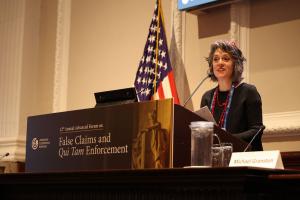 A lady stands on a podium with the American flag in the background at the 12th Annual Advanced Forum on False Claims and Qui Tam Enforcement