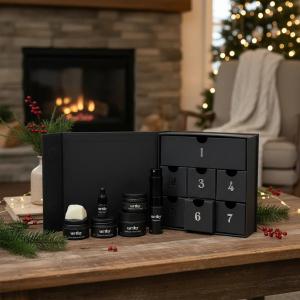 Urthy's 7 Day Advent Calendar gift set displayed on a wooden coffee table in a cozy living room. The open box reveals seven numbered black drawers containing assorted skincare and body products.