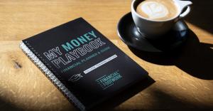 A spiral-bound copy of My Money Playbook: College Training Edition by Financial Footwork sits on a wooden table next to a latte with heart-shaped foam. The financial planner and guidebook is shown in natural sunlight, highlighting its black cover and teal